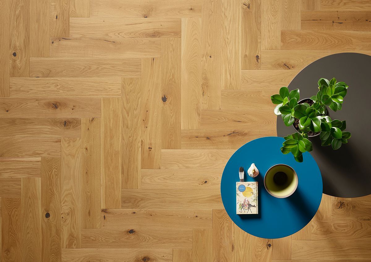 Draufsicht auf einen hellen Holzboden im Fischgrätmuster mit einem blauen runden Tisch, auf dem eine Pflanze und ein kleines Buch stehen.