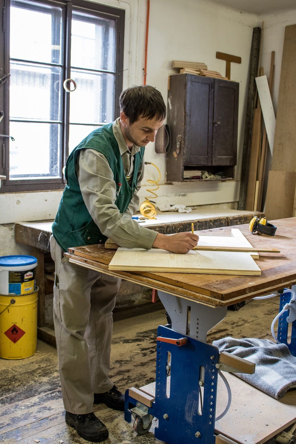 In einer hellen Holzwerkstatt markiert eine Person in einer grünen Weste mit einem Bleistift Maße auf einem Holzbrett an einer Werkbank.