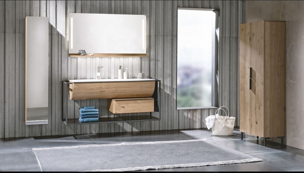 Ein moderner Waschtisch aus hellem Holz mit hohem Schrank, Spiegel und offenen Regalen vor einer grauen Paneelwand.