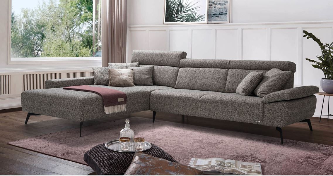 Ein graues, gemustertes Ecksofa mit einer roten Tagesdecke auf einem rosa Teppich in einem hellen, modernen Wohnzimmer.