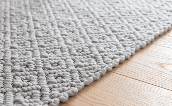 Nahaufnahme eines hellgrauen, strukturierten Webteppichs mit Rautenmuster, der auf einem hellen Holzboden liegt.