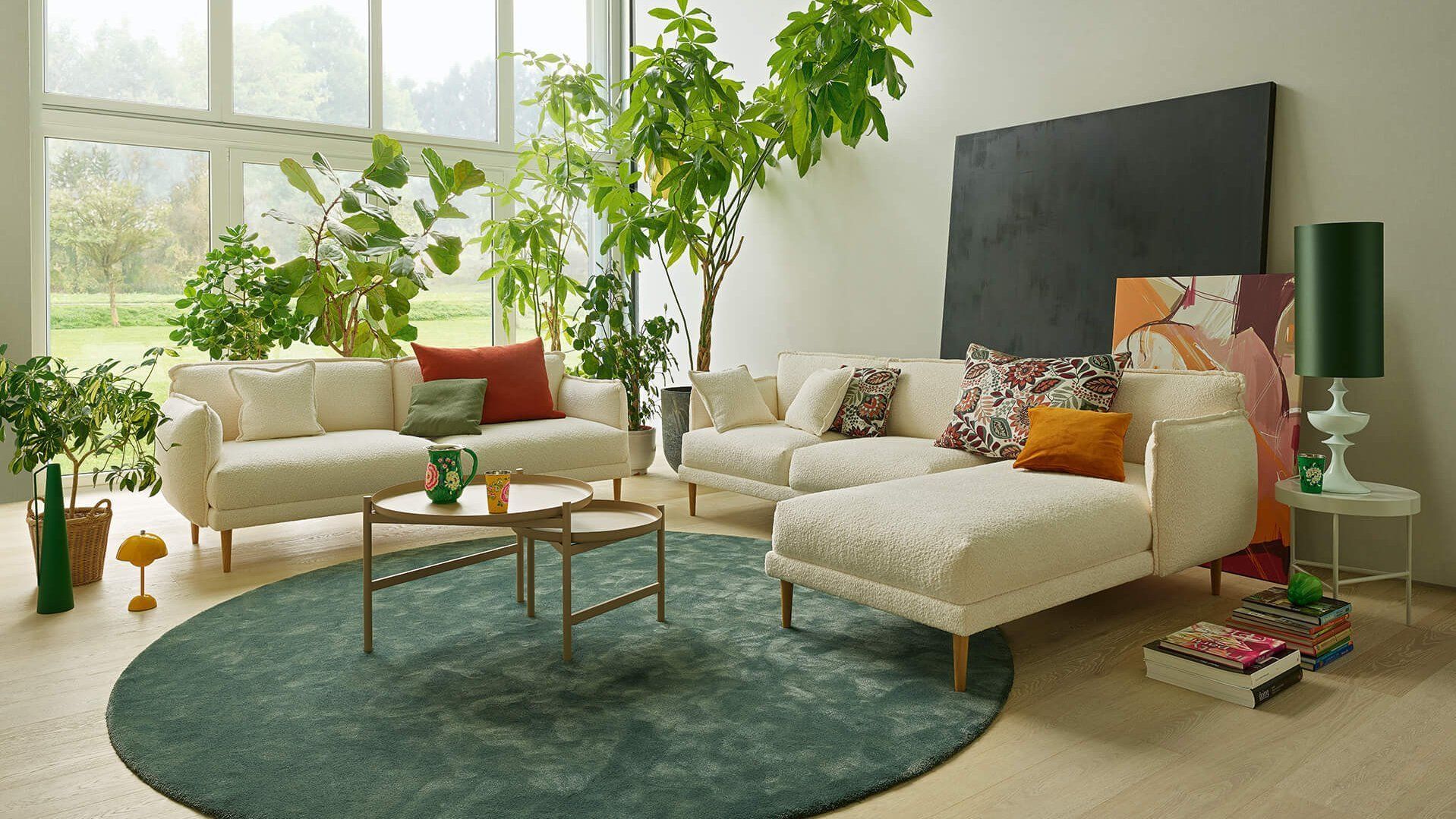 Ein helles Wohnzimmer mit cremefarbenen Sofas auf einem großen runden türkisfarbenen Teppich, umgeben von üppigen Pflanzen und Dekoration.