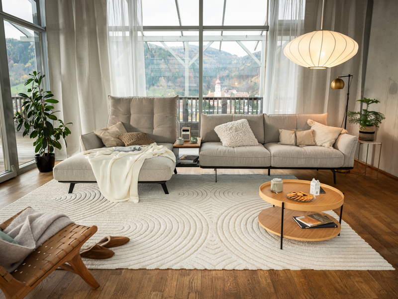 Ein helles, modernes Wohnzimmer mit einem hellgrauen Ecksofa, einem runden Couchtisch aus Holz und einem strukturierten Teppich.