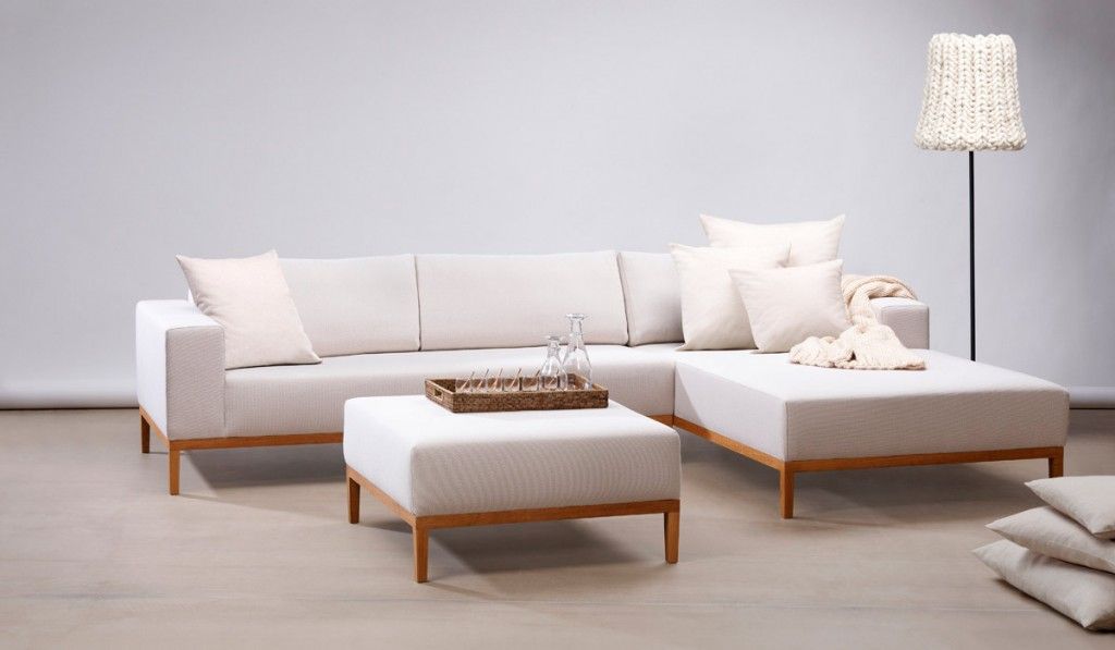 In einem minimalistisch und neutral eingerichteten Studio-Raum stehen ein helles Ecksofa und ein dazu passender Hocker mit Holzbeinen.