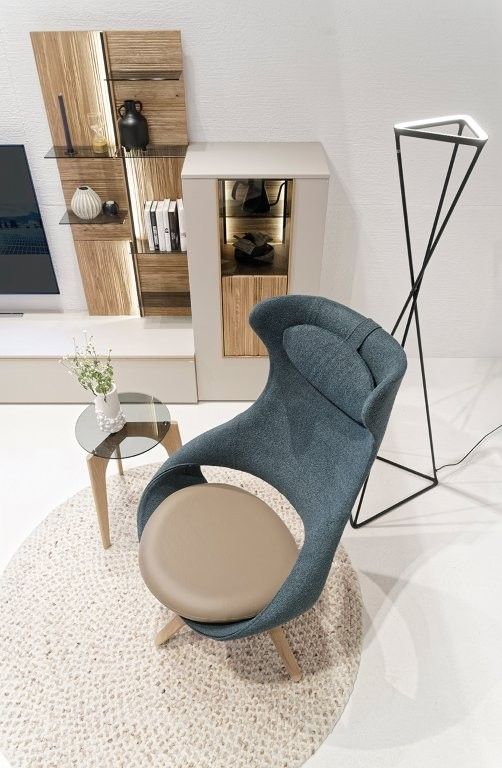 Ein moderner, hochlehniger blauer Sessel mit hellbraunem Sitz steht auf einem runden Teppich neben einem Glastisch und einer geometrischen Stehlampe.