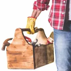 Persona en jeans y camisa a cuadros sosteniendo una caja de herramientas de madera con herramientas, usando un guante amarillo.