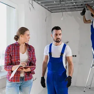 Una mujer con camisa a cuadros y jeans revisa planos con un hombre con overol azul dentro de una habitación en construcción; un trabajador en una escalera instala accesorios de techo.