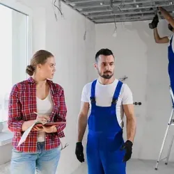 Una mujer con camisa a cuadros y jeans revisa planos con un hombre con overol azul dentro de una habitación en construcción; un trabajador en una escalera instala accesorios de techo.