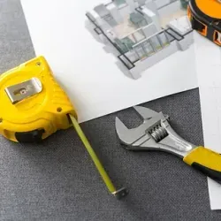 Una cinta métrica amarilla, una llave inglesa ajustable y planos arquitectónicos sobre una superficie gris.
