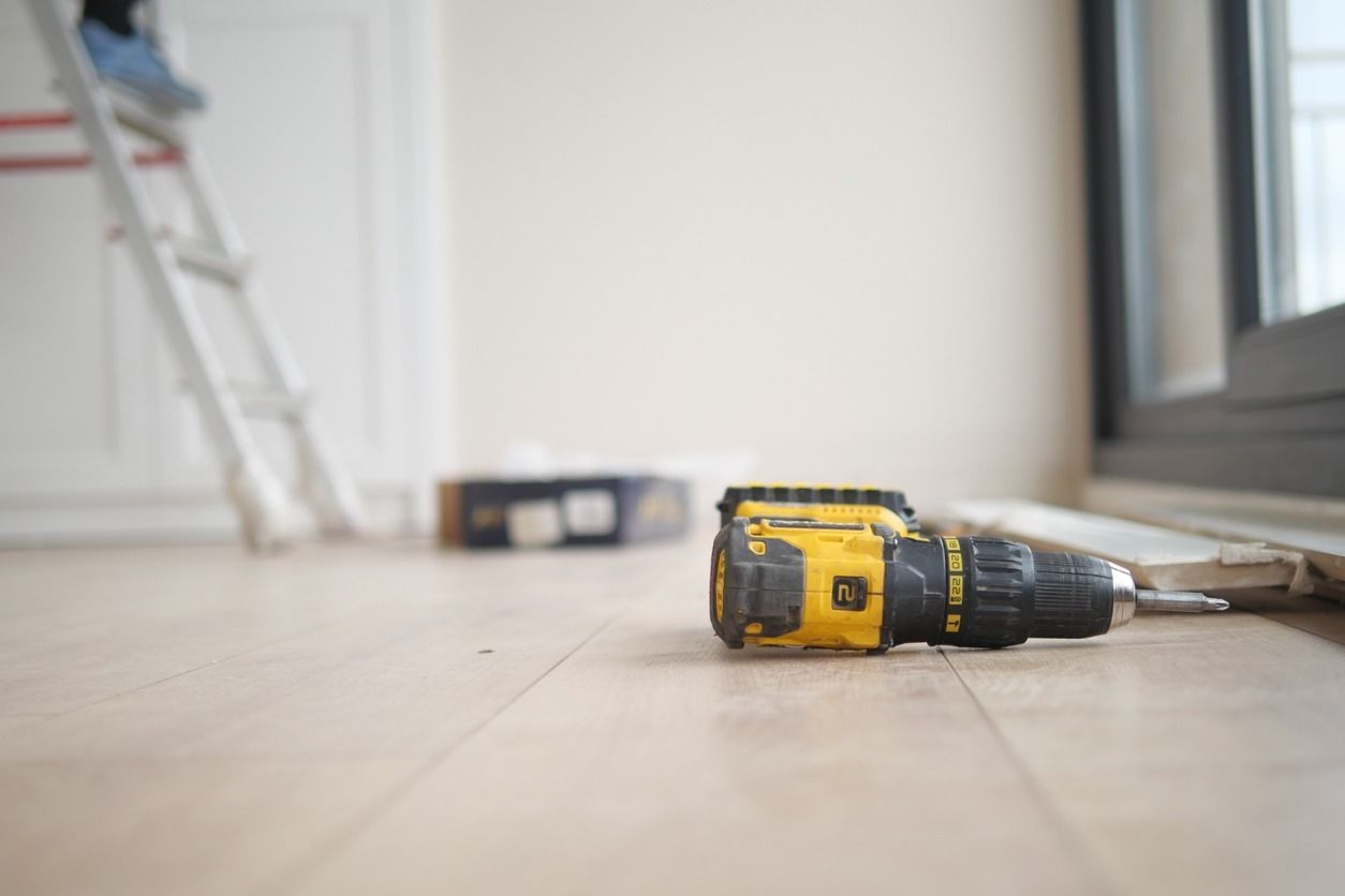 Un taladro eléctrico de color amarillo y negro reposa sobre un piso de madera de color claro, cerca de una escalera y una ventana blancas.