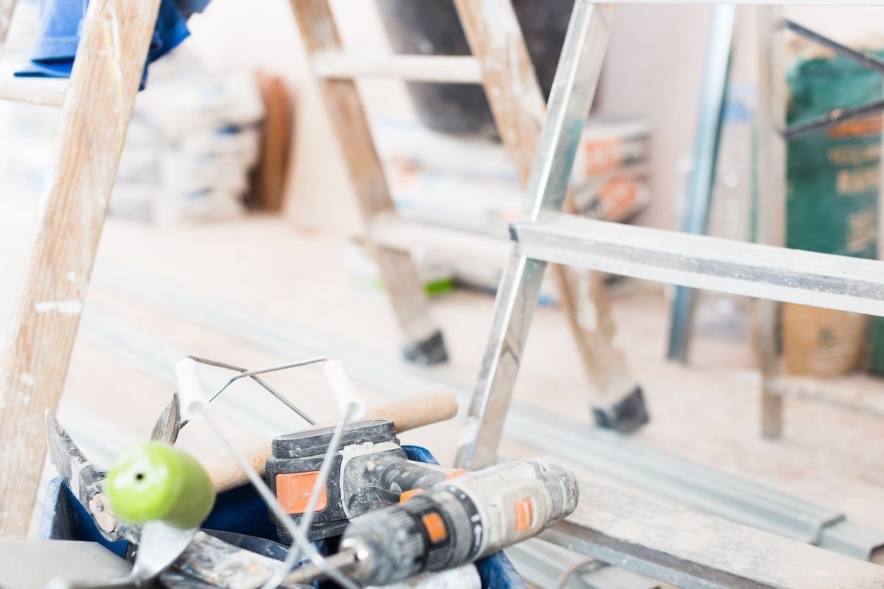 Herramientas y escaleras en un entorno de construcción; un taladro eléctrico y otras herramientas están en foco, con una escalera de madera y una escalera de metal en el fondo.