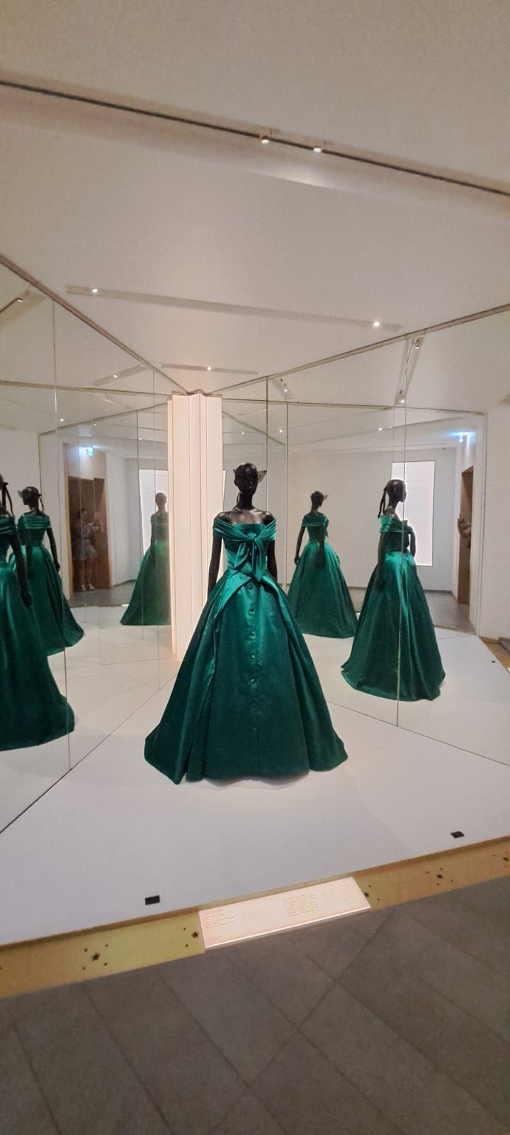 Un mannequin vêtu d'une robe de soirée verte dans une galerie, entouré de miroirs inclinés multipliants son reflet