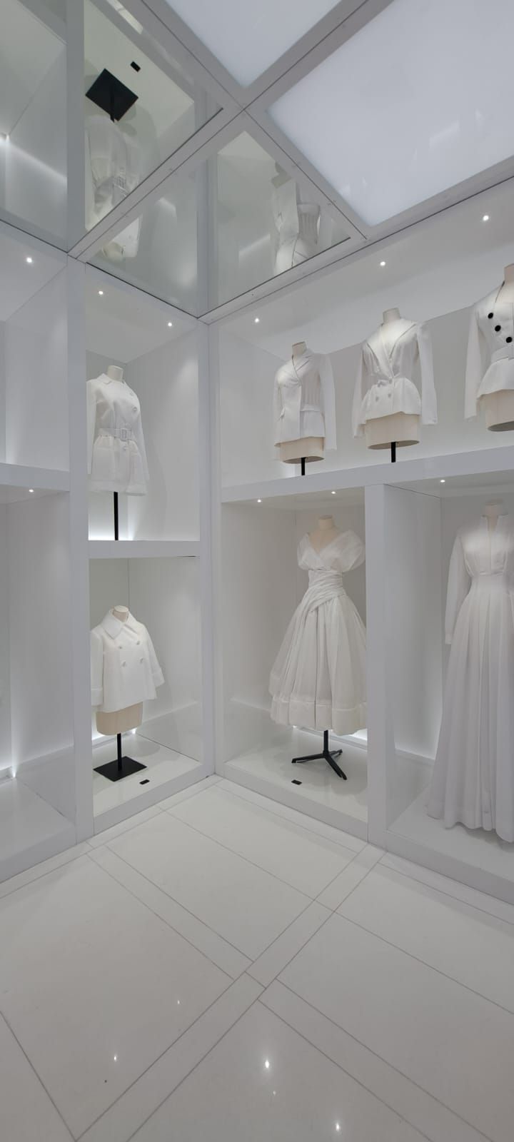 Une vitrine lumineuse et blanche présente plusieurs mannequins vêtus de tenues blanches, disposés sur des étagères
