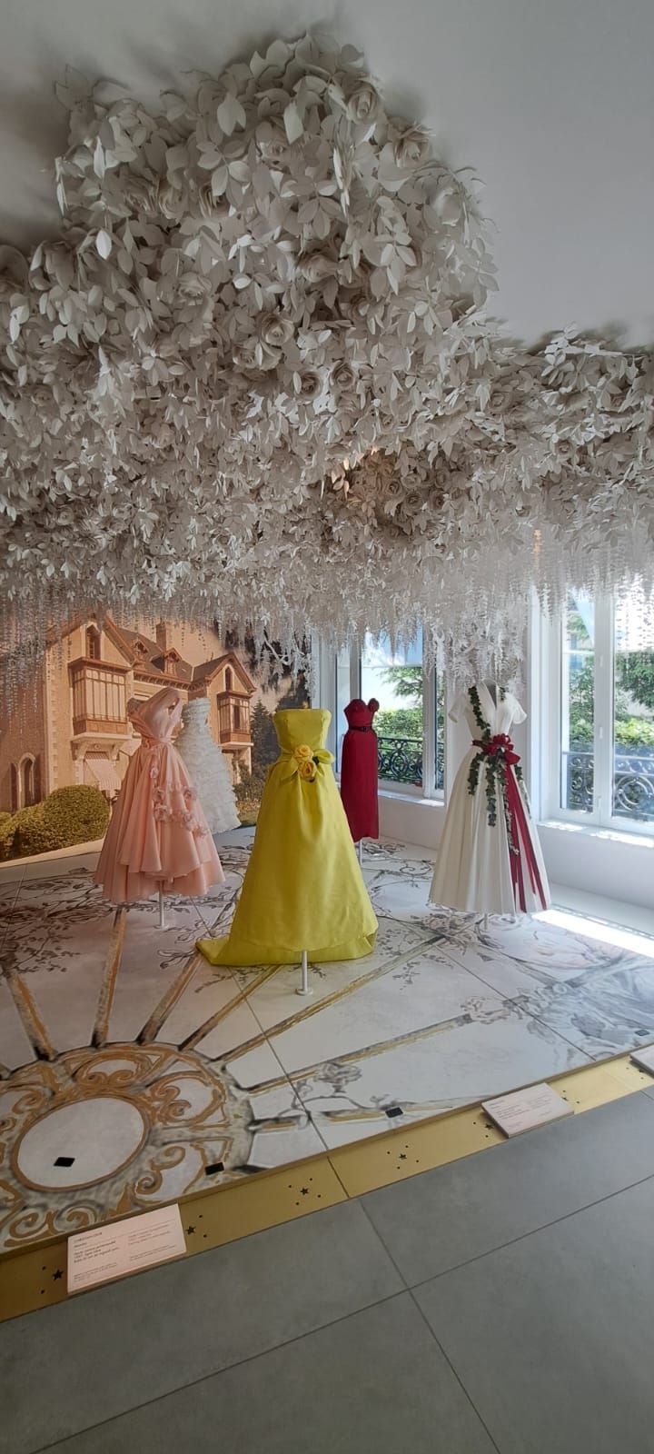 Des robes de couleurs variées sont exposées dans une pièce ornée d'un grand plafond floral travaillé
