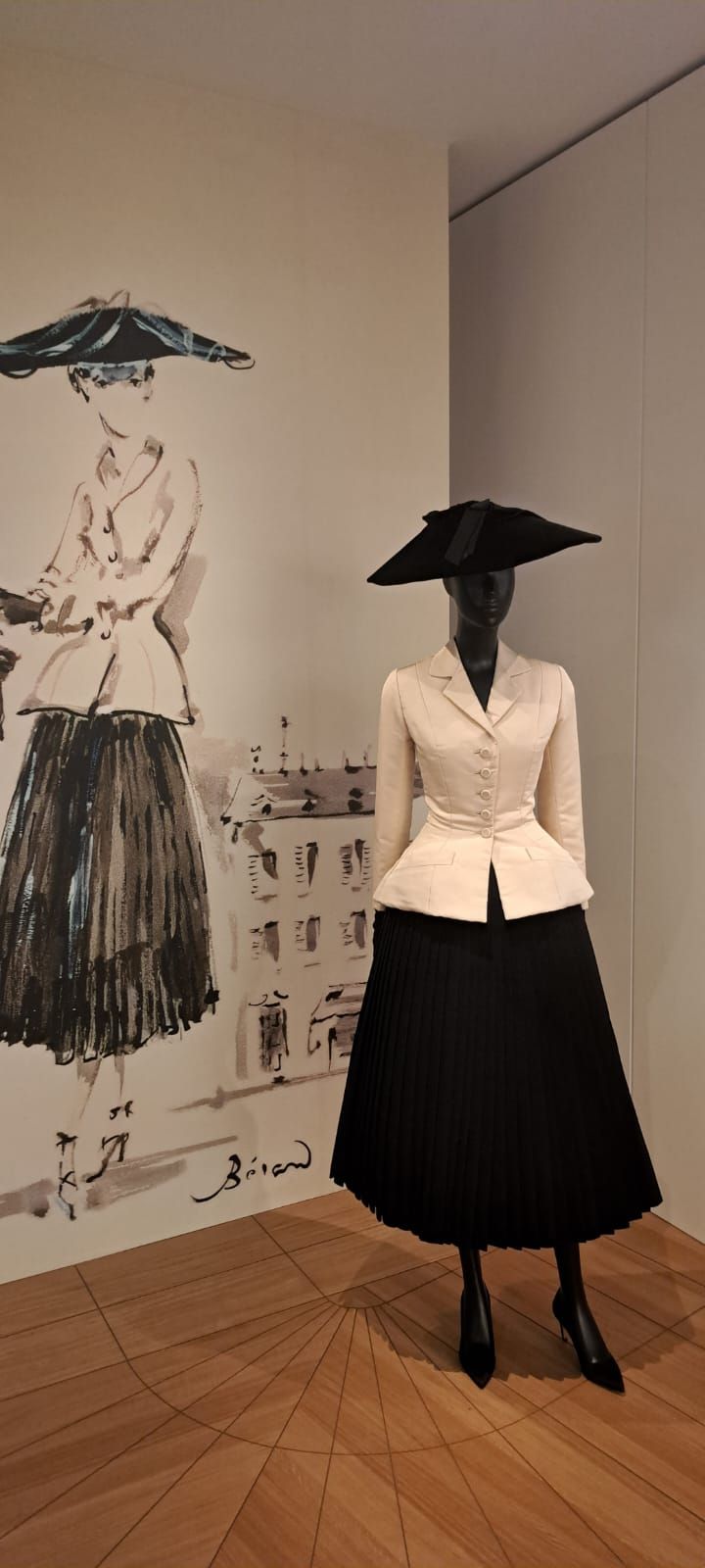 Un mannequin présente une veste blanche et une jupe noire à côté d'un croquis de mode assorti, accroché au mur d'une galerie