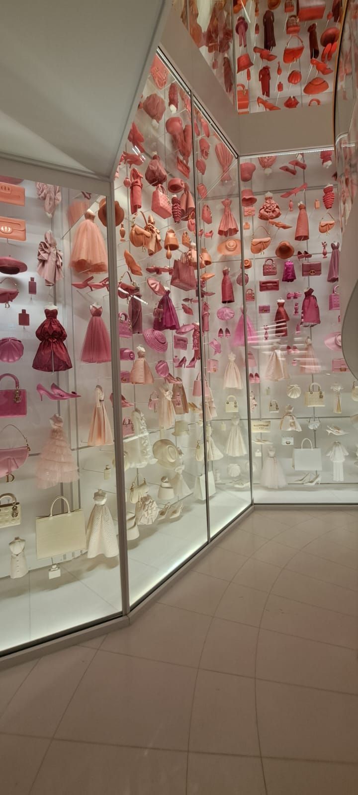 Une vitrine en verre remplie de nombreuses petites robes roses et d'accessoires, disposés par dégradé de couleurs