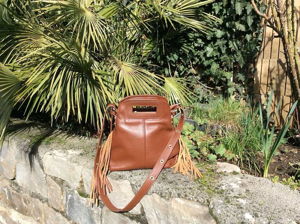 Un sac à main en cuir marron posé sur un muret en pierre dans un jardin, avec des palmes vertes en arrière-plan.