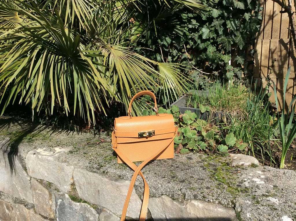 Un sac à main orange posé sur un rebord de pierre dans un jardin luxuriant, avec des feuilles de palmier et de la verdure en arrière-plan.