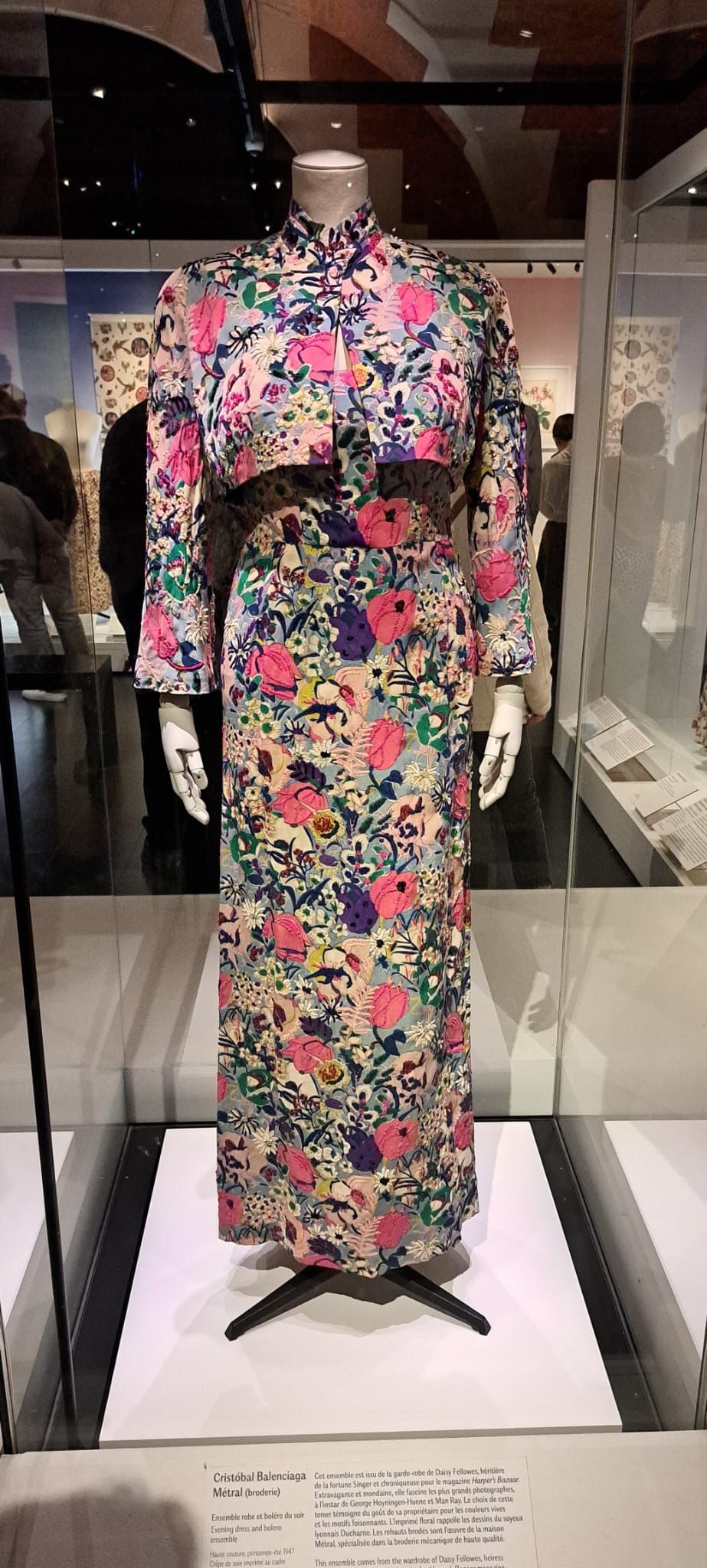 Une robe fleurie colorée à manches longues, ornée de dentelle noire, présentée sur un mannequin dans une vitrine de musée