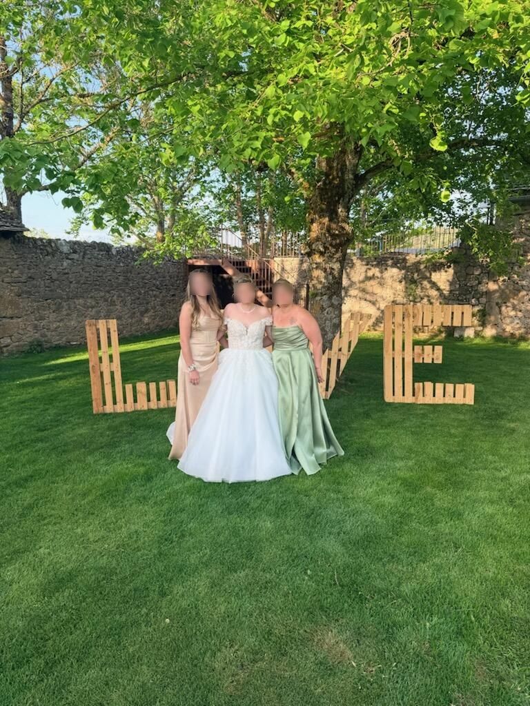 Trois personnes en robes pastel se tiennent sur une pelouse devant de grandes lettres en bois formant le mot « AMOUR ».