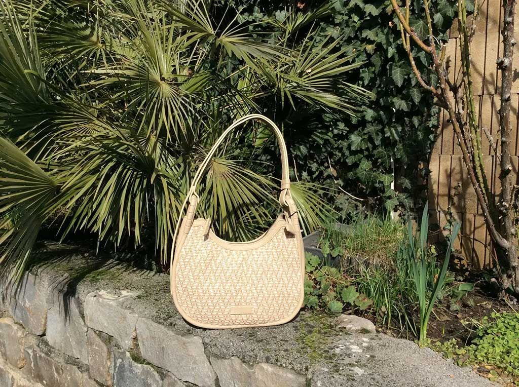 Sac à main en paille tressée avec anse arquée, posé sur un rebord de pierre à côté de plantes verdoyantes.