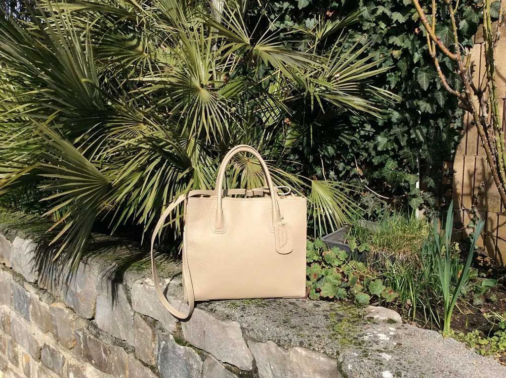 Sac à main beige posé sur un rebord de pierre, à côté de palmiers verts, à l'extérieur.