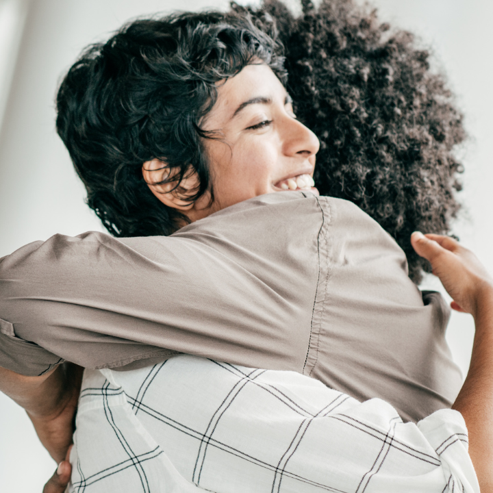 Dos personas abrazándose; una sonriendo, vistiendo una camisa color canela, la otra vistiendo una camisa a cuadros blanca.