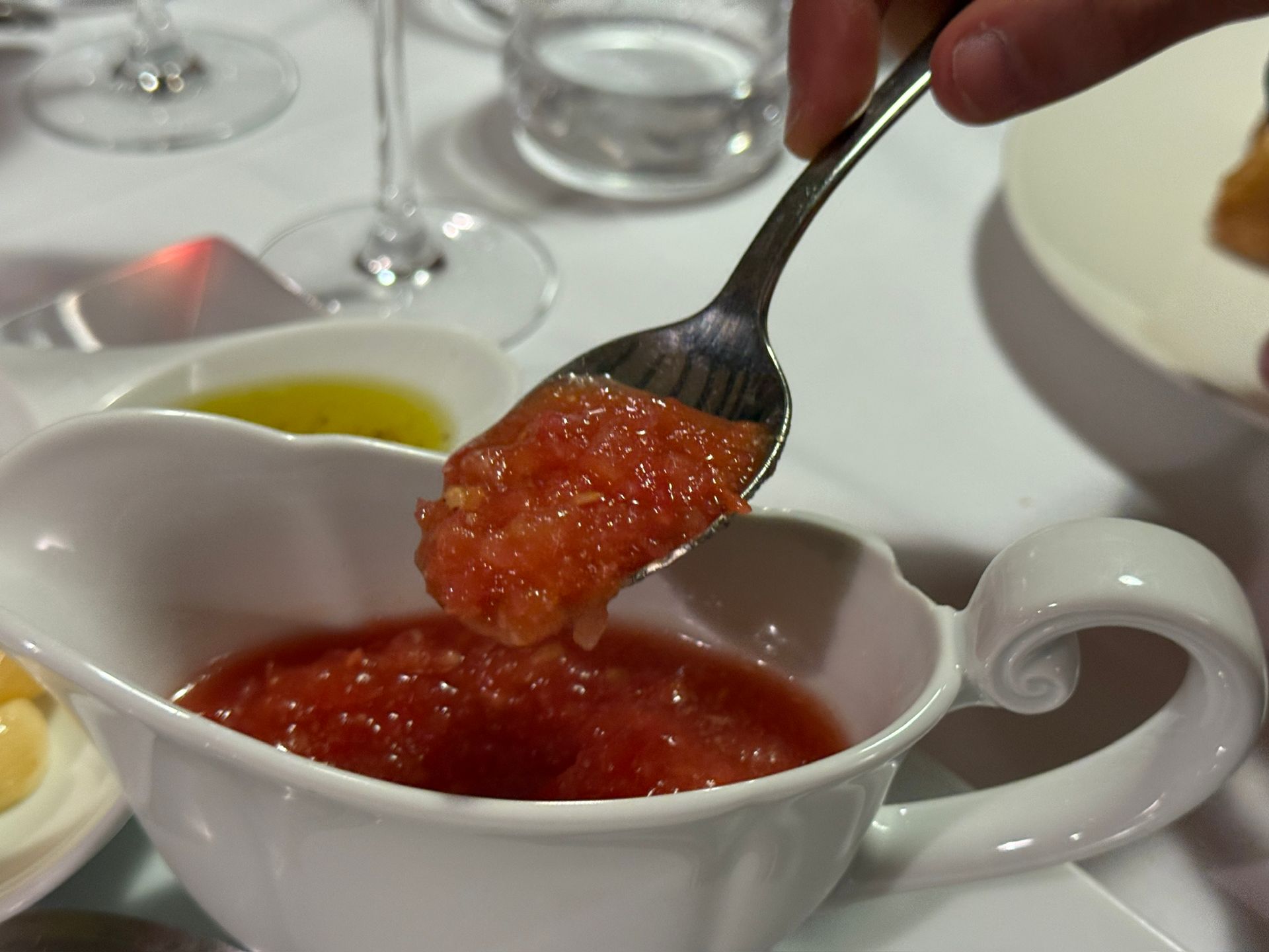 Persona sosteniendo una cuchara de salsa de tomate triturada sobre una salsera blanca; mantel blanco.