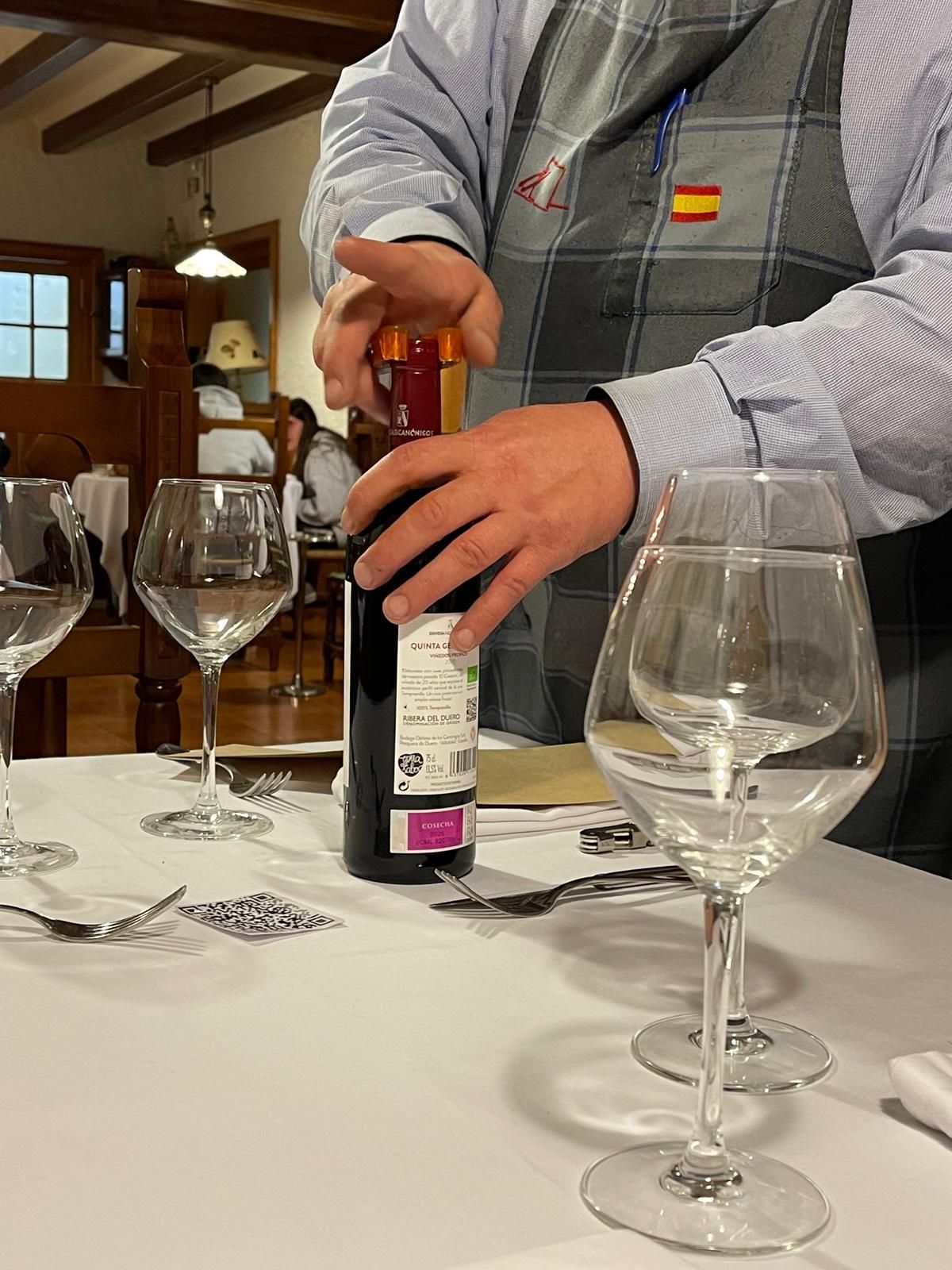 Persona abriendo una botella de vino en una mesa puesta con copas de vino en un restaurante.