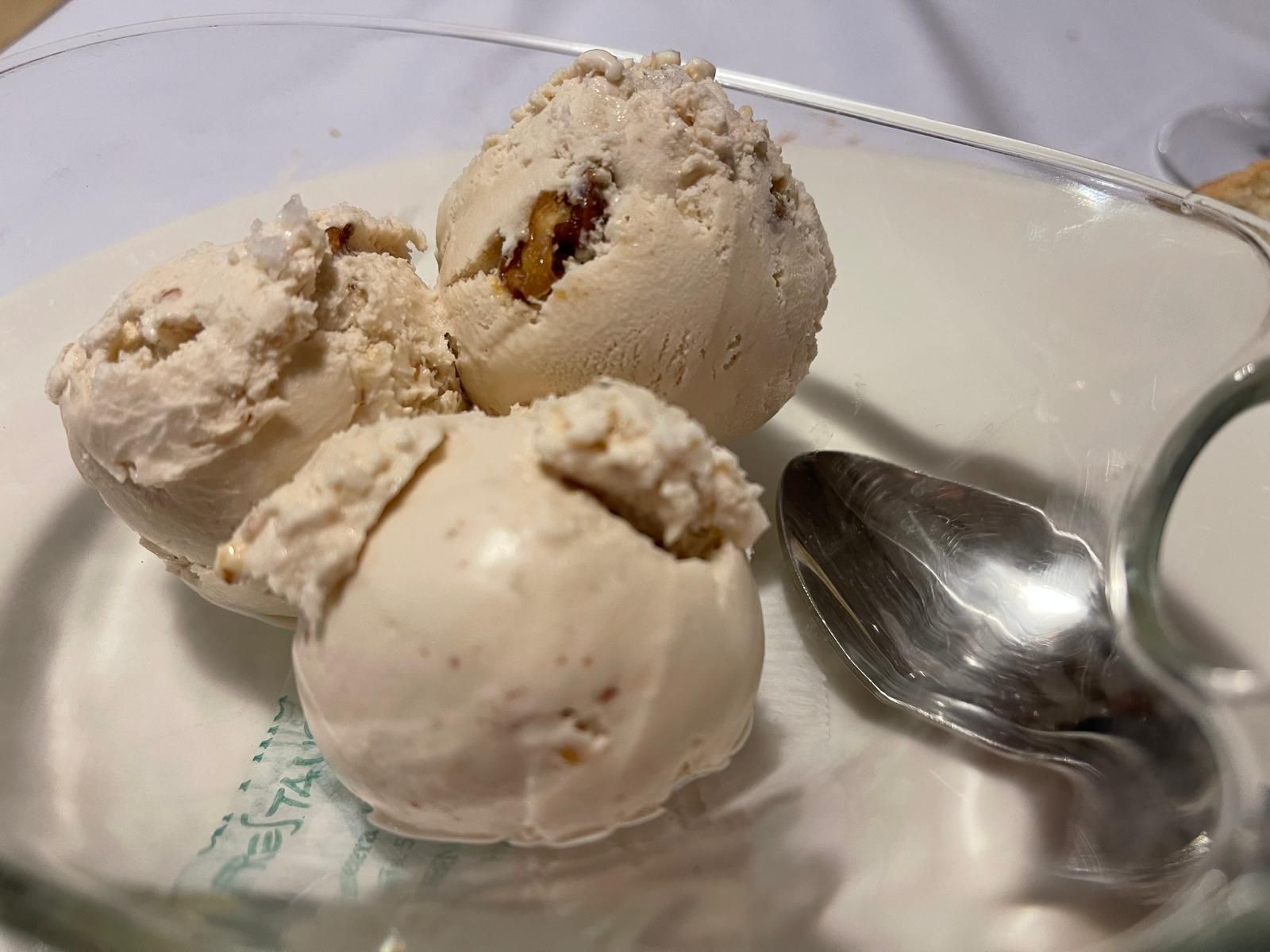 Tres bolas de helado en un plato de vidrio transparente con una cuchara, nueces visibles, color marrón claro.