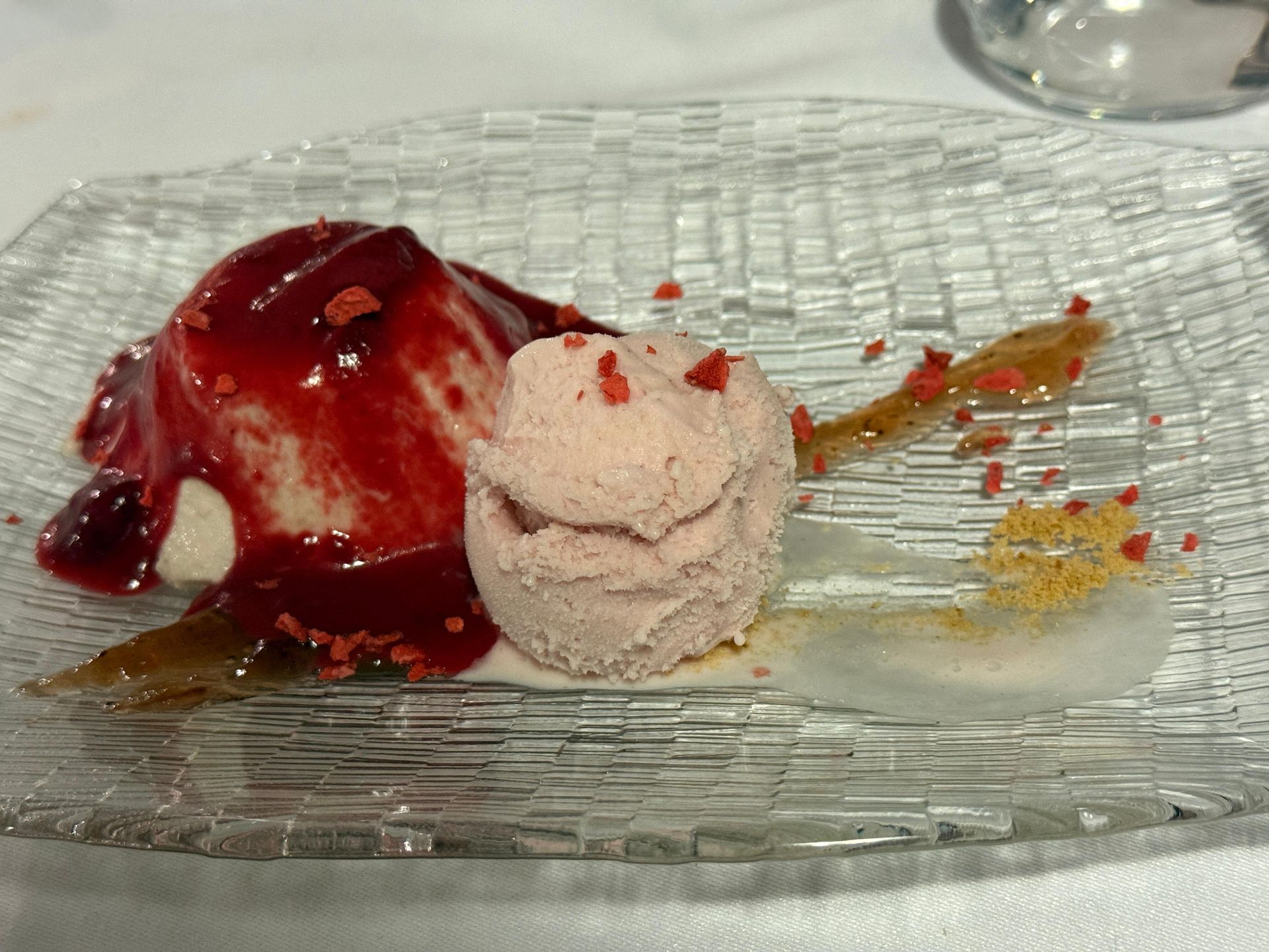 Postre: bola de helado rosa, crema blanca con salsa de frutos rojos y guarnición en plato de vidrio texturizado.