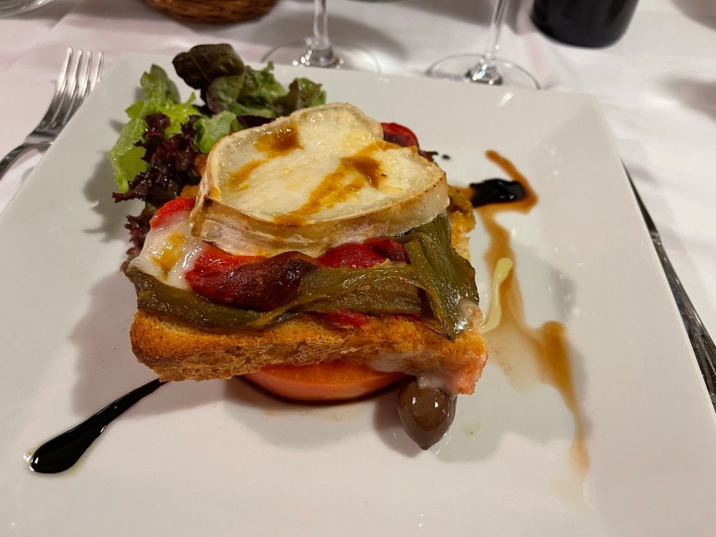 Plato cuadrado con entrante en capas: tomate, tostadas, pimientos, queso fundido y verduras.