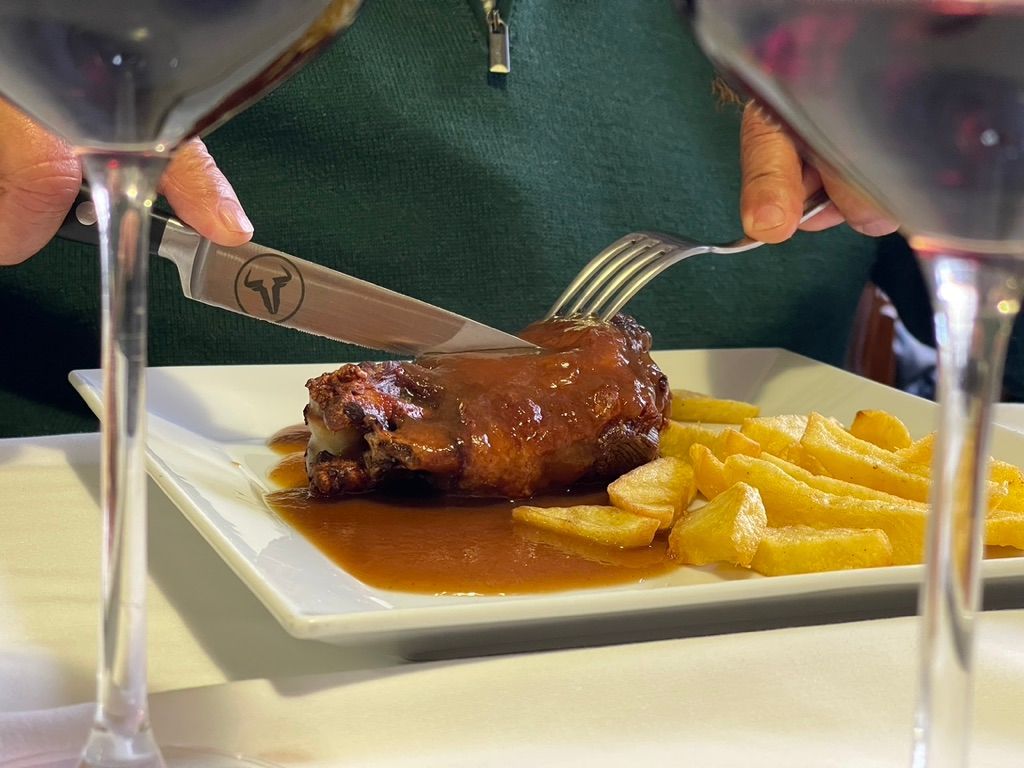 Persona cortando un plato de carne, servido con patatas fritas y salsa, enmarcado por dos copas de vino.