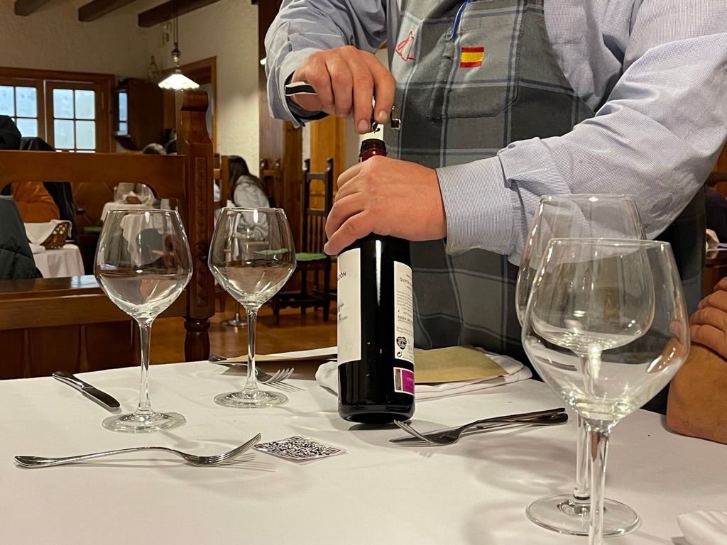 Un camarero abre una botella de vino tinto en una mesa de restaurante con copas de vino dispuestas.