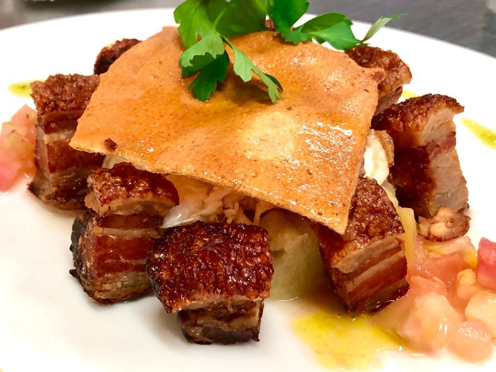 Panceta de cerdo crujiente con galleta y tomates, adornada con perejil en un plato blanco.