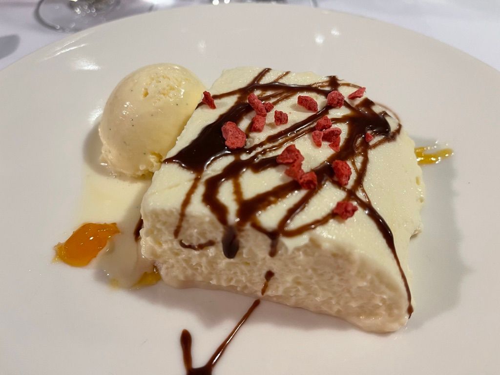 Postre en plato blanco: pastel blanco rociado con chocolate, cubierto con trozos rojos, helado de vainilla.