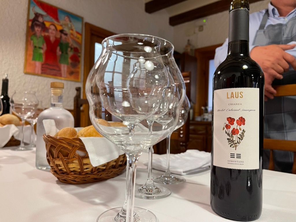 Copas y una botella de vino sobre una mesa. Una persona junto a la botella. Al fondo, una cesta de pan y un cuadro.