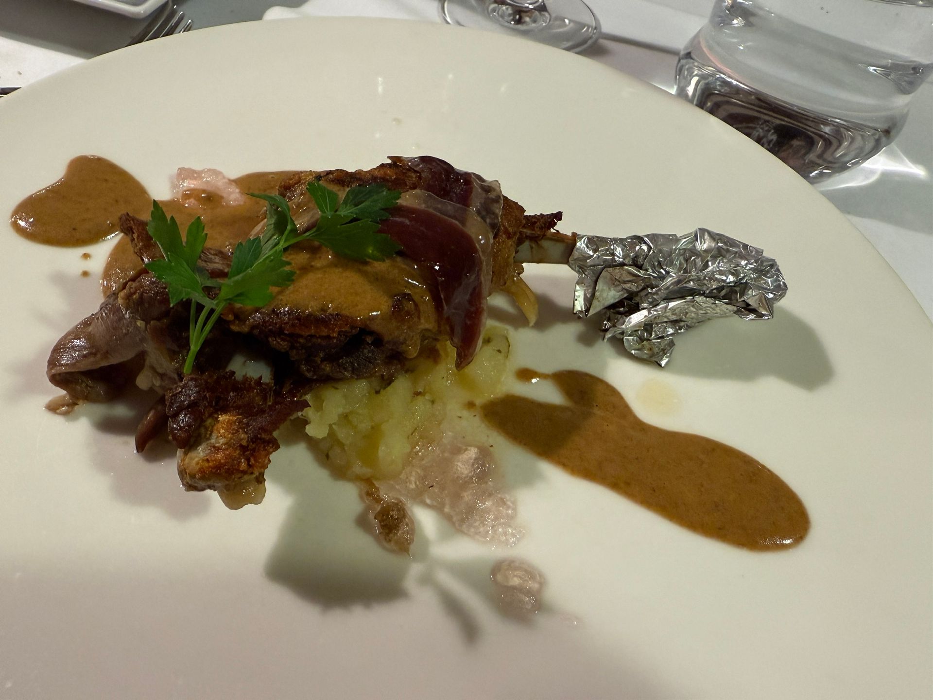 Confit de pato con salsa en un plato blanco, adornado con perejil. Hueso cubierto con papel de aluminio.