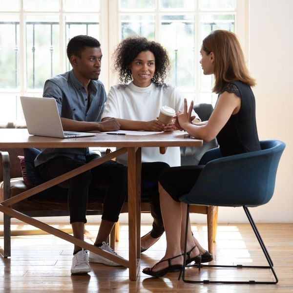 Tres personas en una oficina luminosa, reunidas alrededor de una mesa de madera, conversando con una computadora portátil abierta.