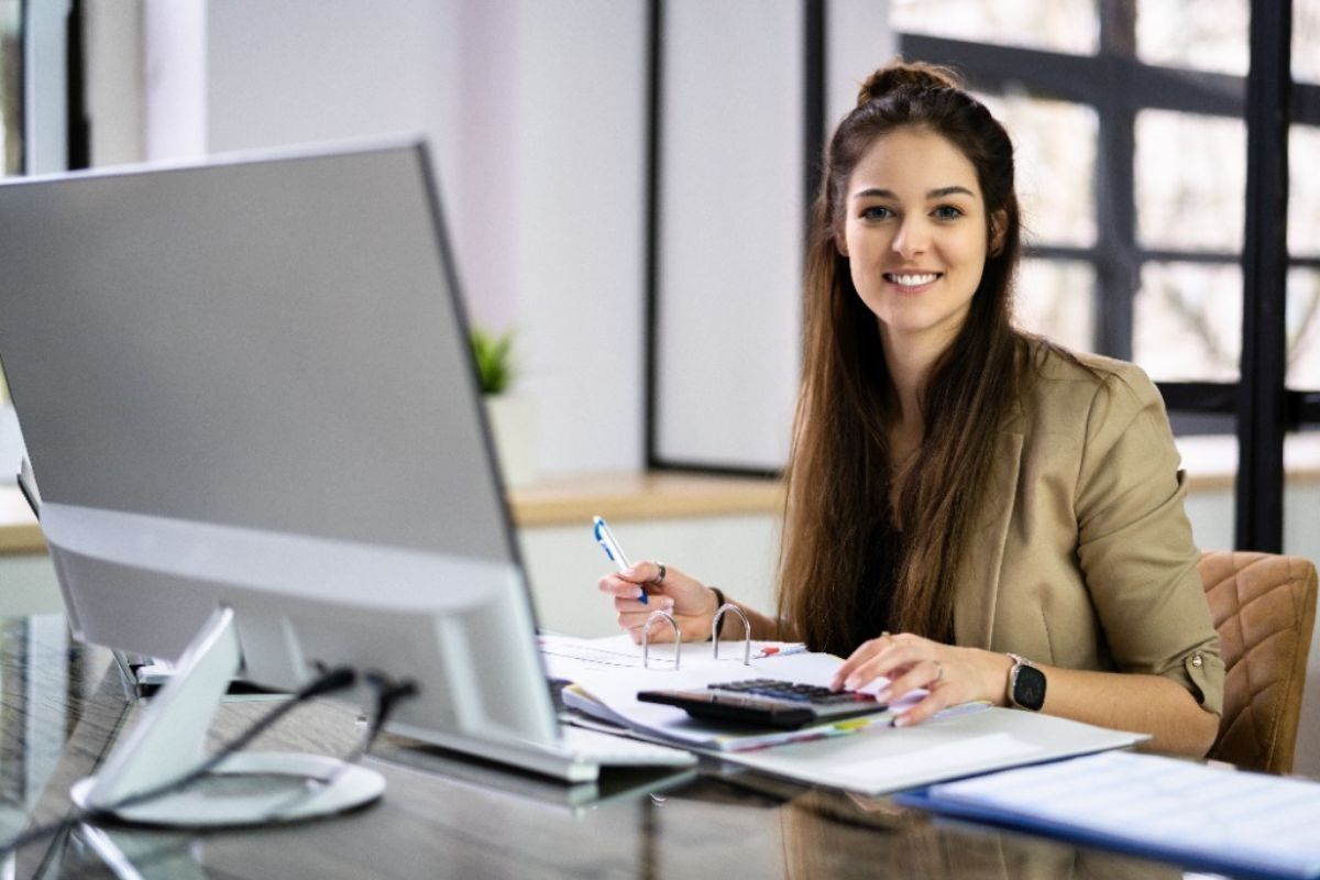 Mujer sonriente sentada en un escritorio de una oficina, sosteniendo un bolígrafo junto a un monitor de computadora y papeles.