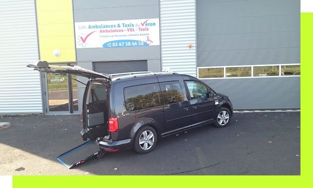 Fourgonnette gris foncé accessible aux fauteuils roulants avec rampe déployée, stationnée devant un bâtiment.