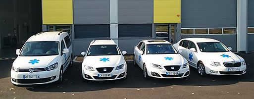 Quatre véhicules blancs arborant des symboles médicaux bleus sont stationnés devant un bâtiment gris et jaune.