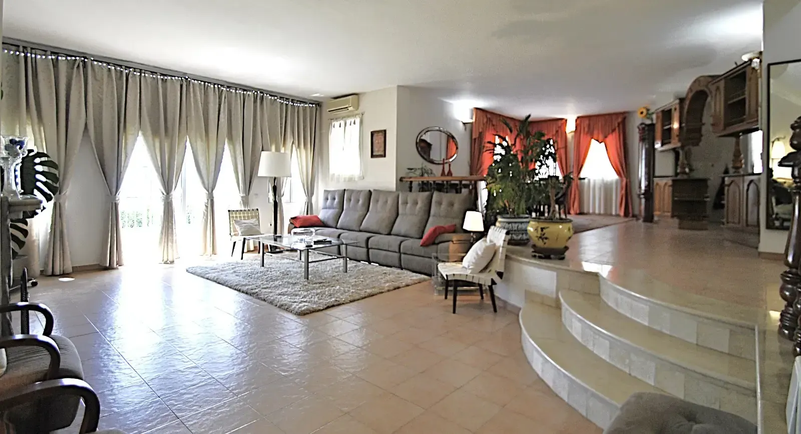 Sala de estar luminosa con grandes cortinas, sofás grises, suelo de baldosas y una escalera blanca curva.