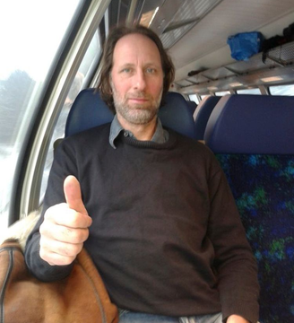 Mann im Zug zeigt den Daumen nach oben; er hat braunes Haar und einen Bart und trägt einen schwarzen Pullover.