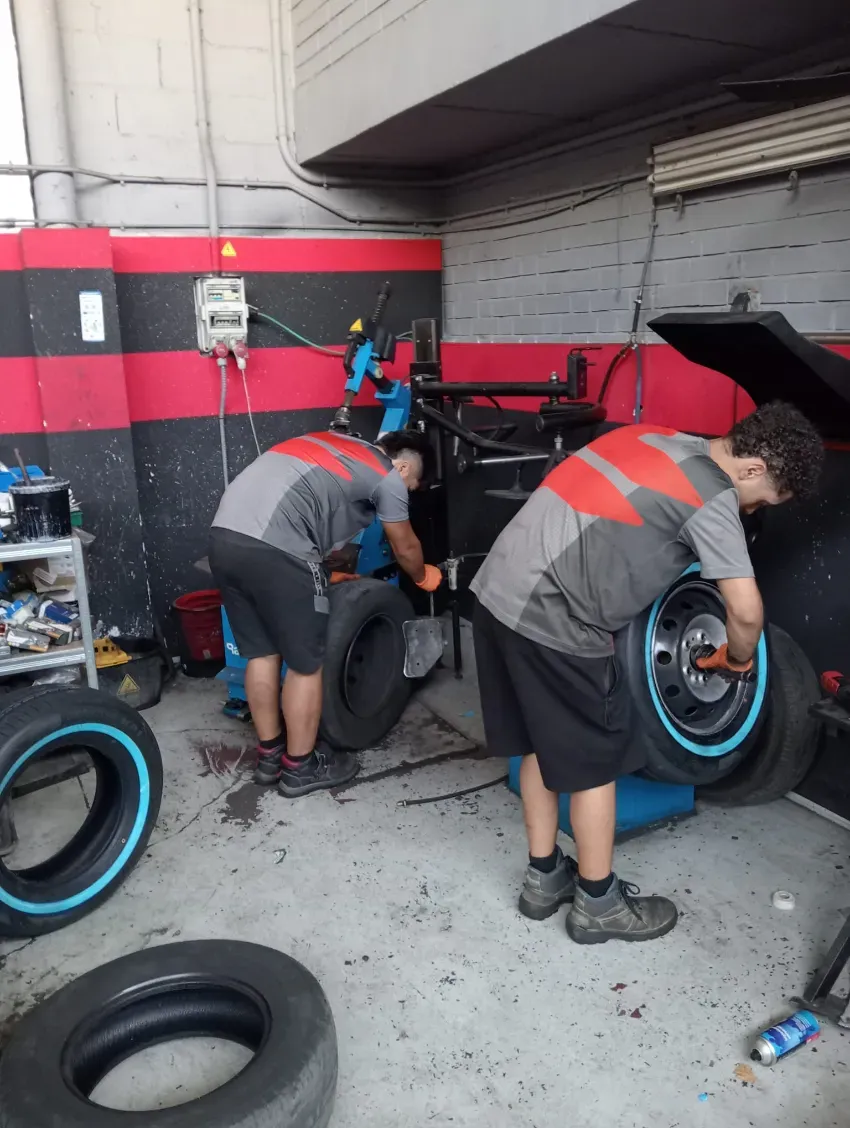 Dos mecánicos cambiando un neumático de motocicleta en un taller rojo y negro, con ruedas y herramientas en el suelo.