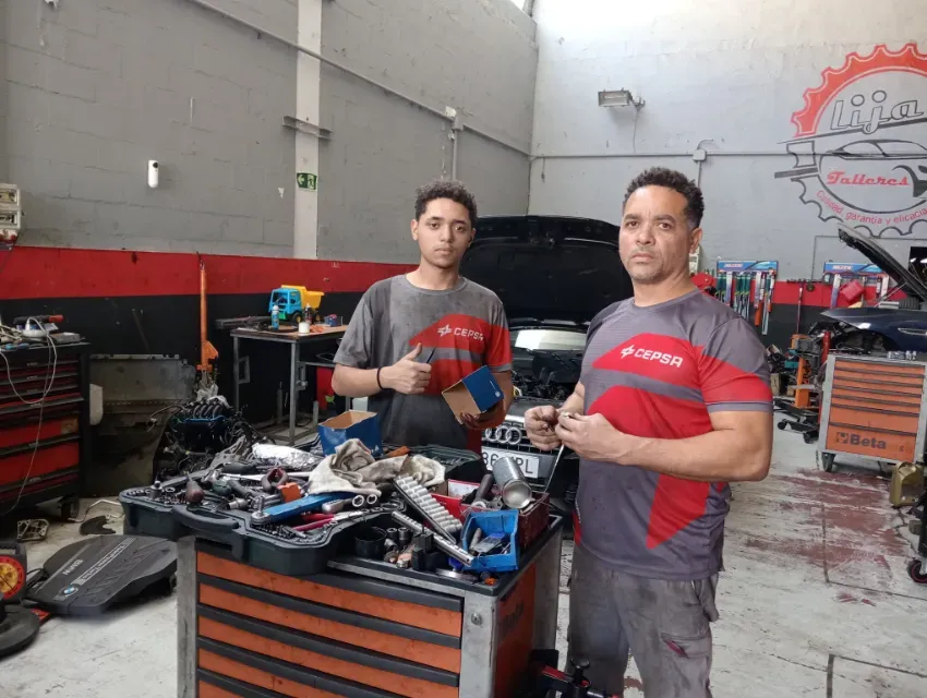 Dos mecánicos en un taller sosteniendo piezas rojas de automóviles junto a un carro de herramientas.