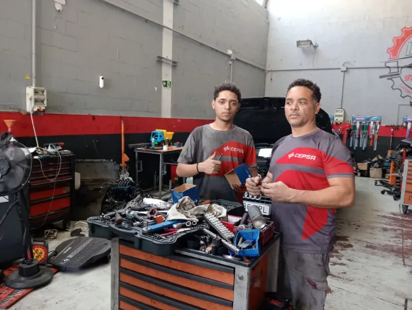 Dos mecánicos en un taller, de pie junto a un banco de trabajo cubierto de herramientas y piezas de motor.