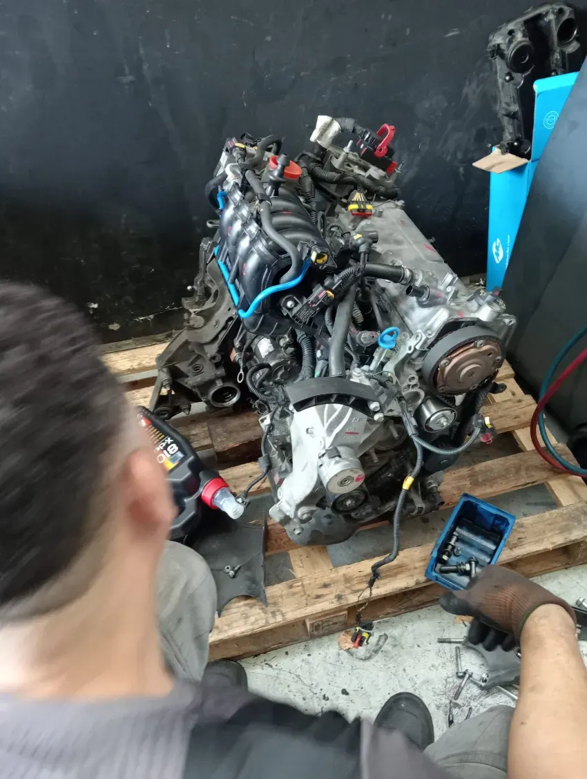 Un mecánico trabaja en el motor de un coche sobre un palé en un garaje, con herramientas a su alcance.