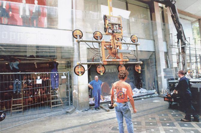 Ein Kran hebt eine große Glasscheibe zum Einbau an, Bauarbeiter beobachten das Geschehen.