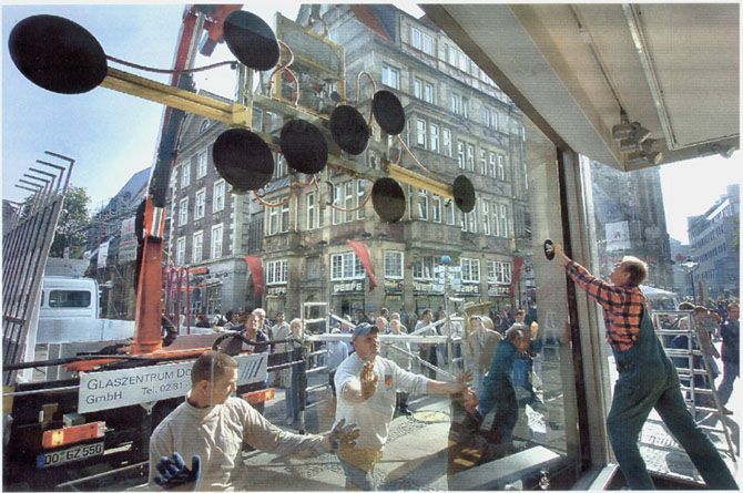 Arbeiter montieren mit einem Kran eine große Glasfensterscheibe. Straßenszene, im Hintergrund ein mehrstöckiges Gebäude.